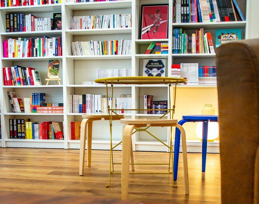 Intérieur de la librairie Le vestiaire à mot à Nice 
Au premier plan, une table avec trois tabouret posée devant une étagère de livres