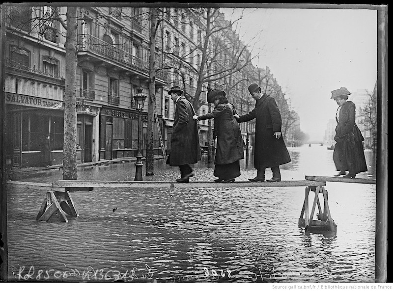 Photo en noir et blanc de 4 personnes traversant une route inondée sur des planches posées sur des tréteaux