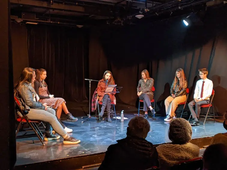 Plusieurs personnes sont sur scène. L'une d'elles parle dans un micro en lisant un texte.