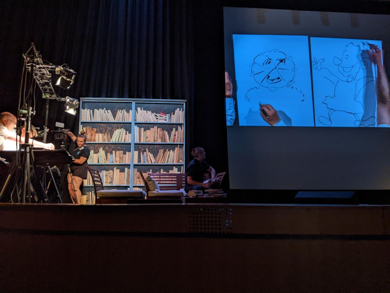 Sur une scène, deux auteurs dessinent sur une table à dessin. Leurs dessins sont reproduits sur un grand écran.