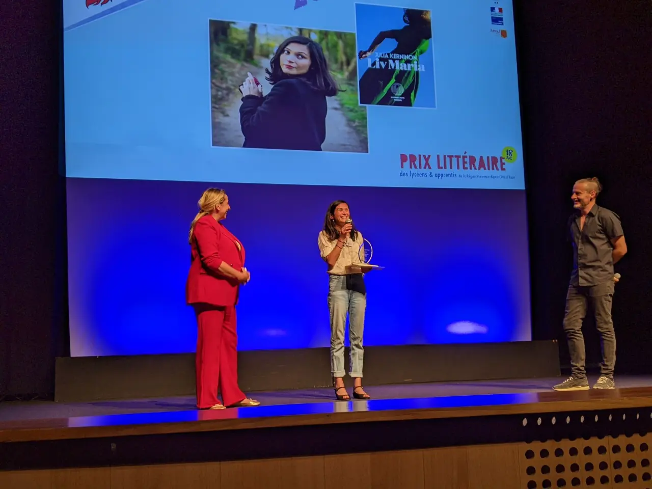 Trois personnes sont sur scène, l'un d'entre elles parle dans un micro. Elle tient un trophée à la main.