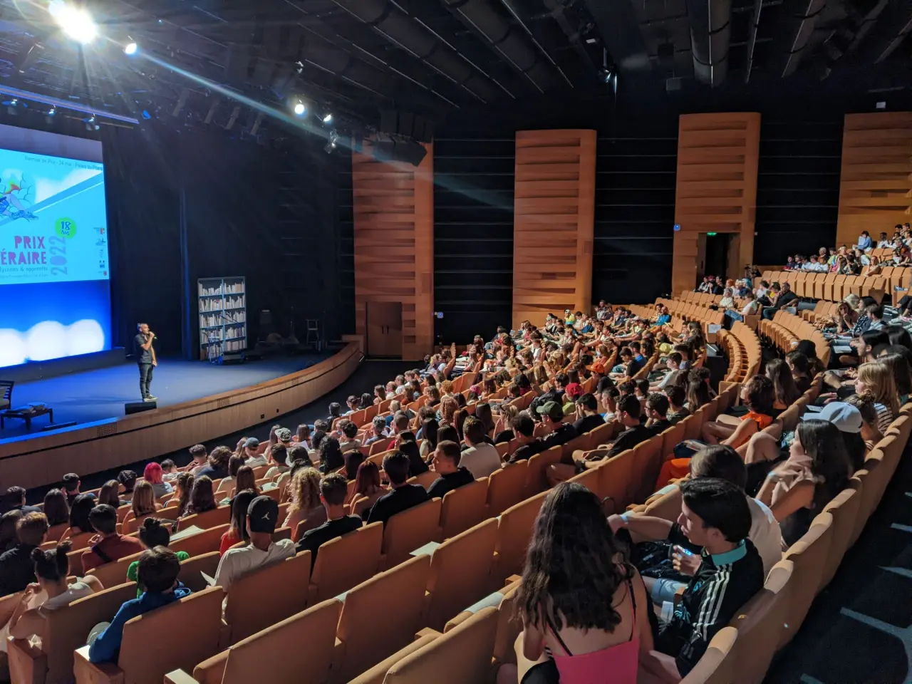 Des lycéens sont assis face à la scène du Palais du Pharo à Marseille. Il y a un animateur sur la scène.