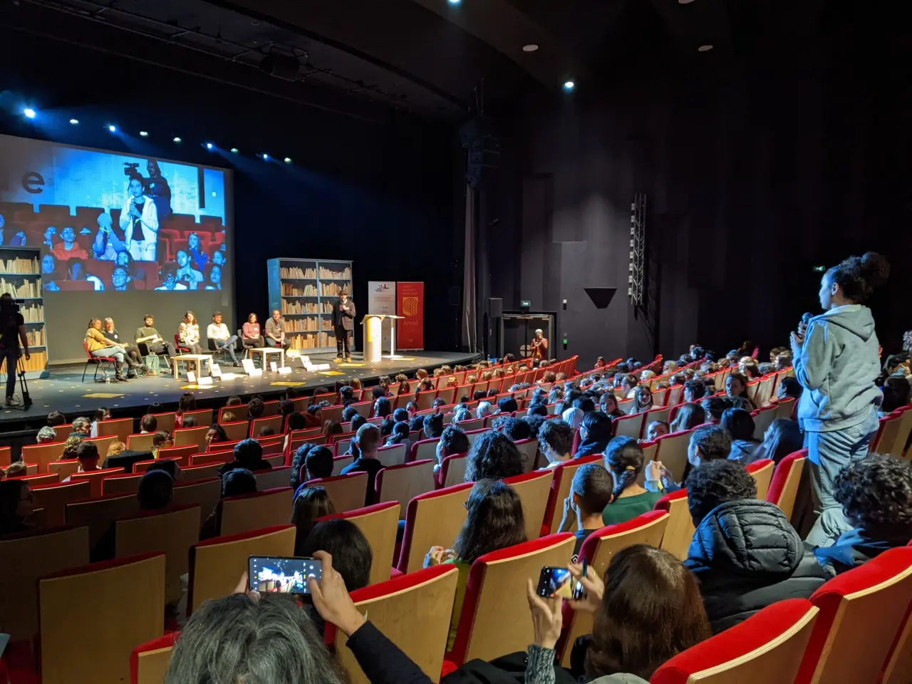 Des lycéens et apprentis sont assis dans un théâtre face à six auteurs. Une élève est debout, elle tient un micro.