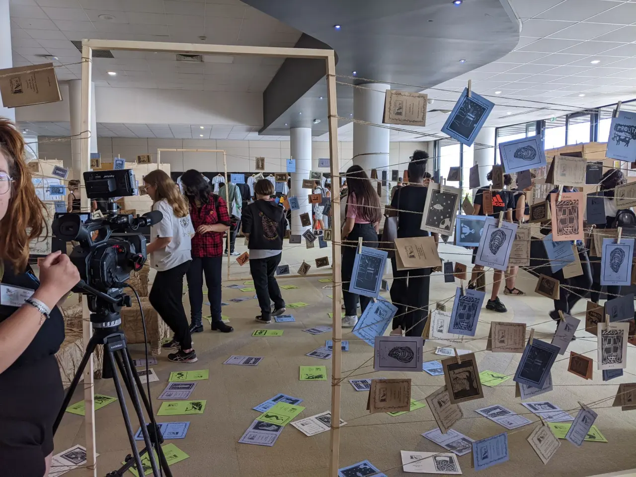 Des lino-gravures sont suspendues avec des épingles sur des cordes. Plusieurs personnes se baladent dans cette exposition en volume.